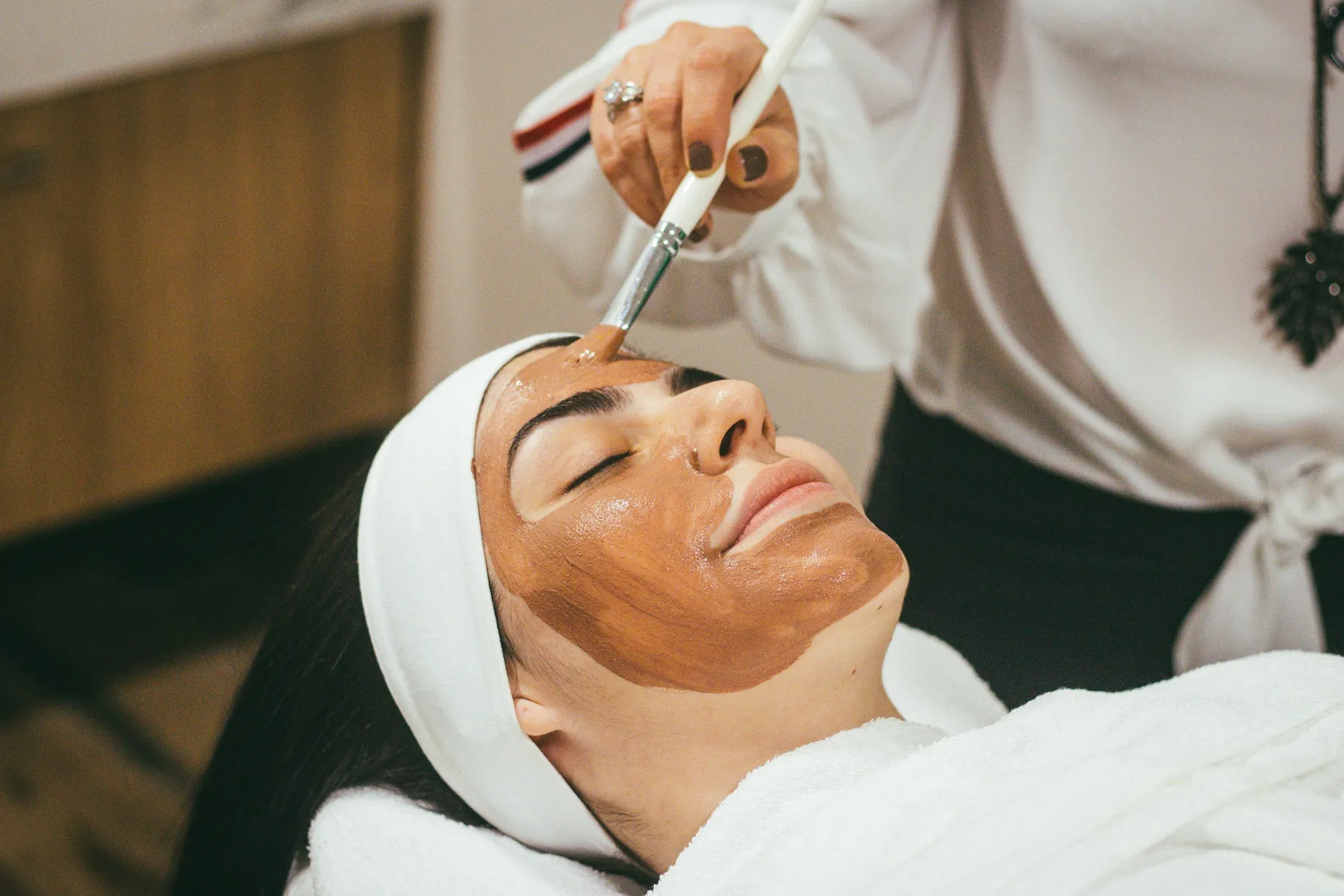 Esteticista aplica máscara facial com pincel em cliente deitada na maca